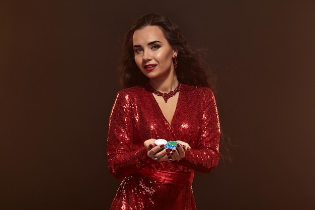 Photo of young beautiful sexy lucky brunette woman in a red evening shiny dress holds poker chips in her hands, showing her winnings. Brown smoky background, gambling industry