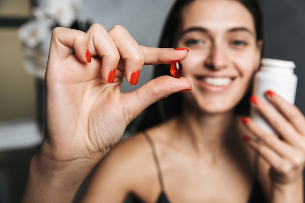 Photo of young beautiful woman in bathroom holding vitamins pills in hands.