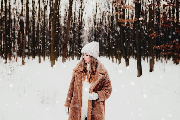 Photo of young beautiful woman in fur coat standing on the white snow in winter forest. Girl is smiling and happy with closed eyes. Female has winter vacation, happily waiting for Christmas holidays