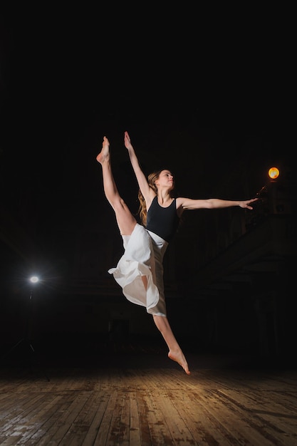 Photo of young brunette dancer girl ballerina in white skirt in split jump on stage