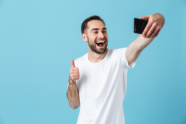 笑って、青で隔離のスマートフォンでselfieを取る基本的なTシャツを着ている若いブルネットの男性の写真