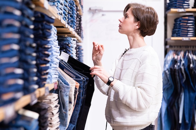 Photo of young brunette woman with a short haircut in a white sweater chooses stylish and casual jeans and denim clothes in a store. Shopping concept