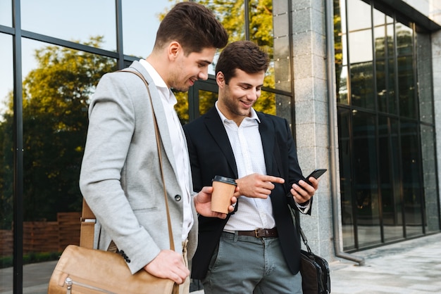 Photo of young businessmen in suits using smartphones, while standing outdoor near building with takeaway coffee