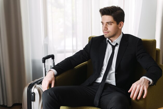 Photo of young calming businessman wearing black suit looking aside and sitting on armchair with suitcase in hotel apartment