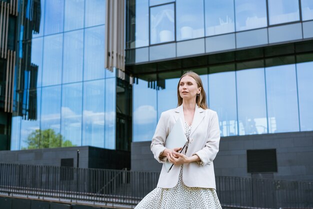 オフィスビルの背景に立っている若い白人の陽気なビジネス女性の写真...