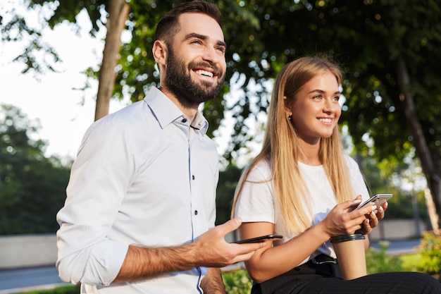 Photo of a young cheerful happy man and woman businesspeople outside in nature park using mobile phones.