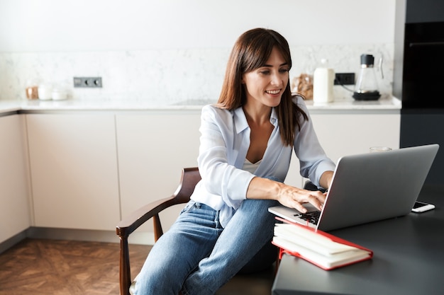 Photo of a young cheerful smiling woman indoors at home at the kitchen using laptop computer.