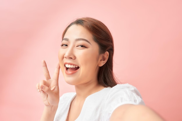 Photo of young cheerful woman happy positive smile make selfie show peace cool vsign isolated over pink background