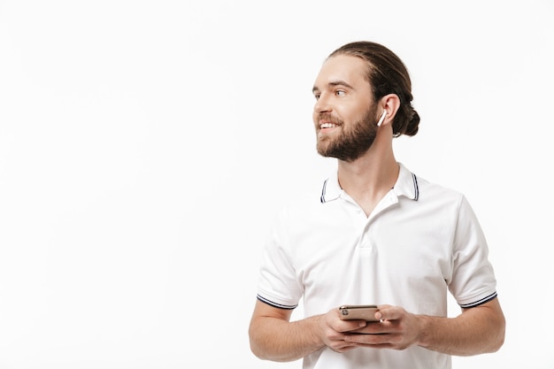 Photo of a young cheery positive happy handsome bearded man posing isolated over white wall using mobile phone listening music with earphones.