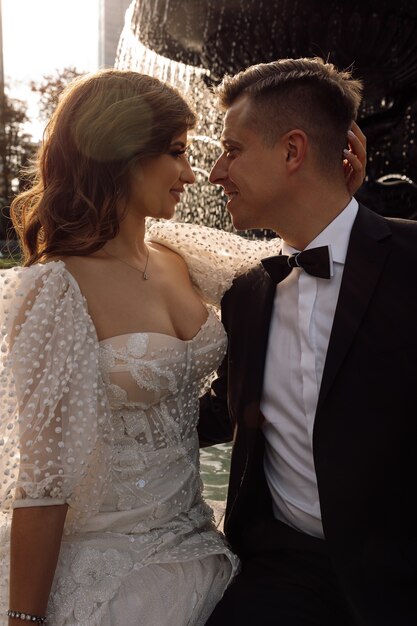 Photo of young couple in love embracing in rays of setting sun against backdrop of urban landscape with fountain, snuggled up to each other, wedding day concept