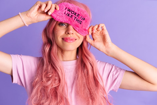 Photo of young cute cheery girl with pink anime hair posing isolted over purple wall with sleeping mask.