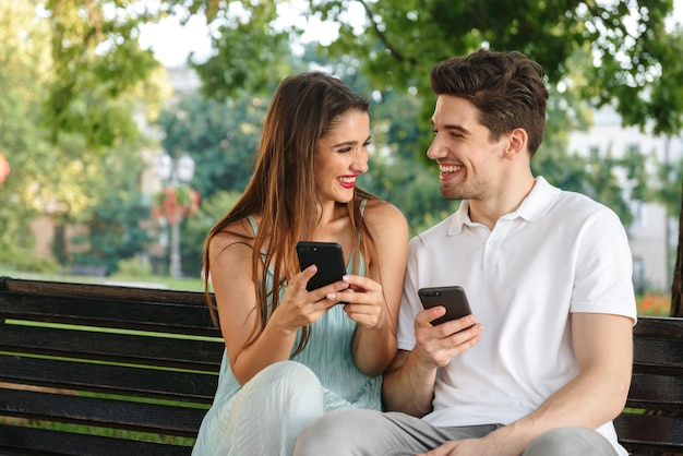 Photo of young cute loving couple sitting outdoors while using mobile phones