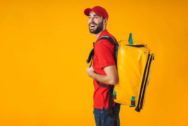 Photo of young delivery man in red uniform carrying backpack with takeaway food isolated over yellow