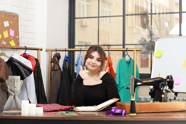 Photo of young female tailor looking at sketch book and gesture thumb up