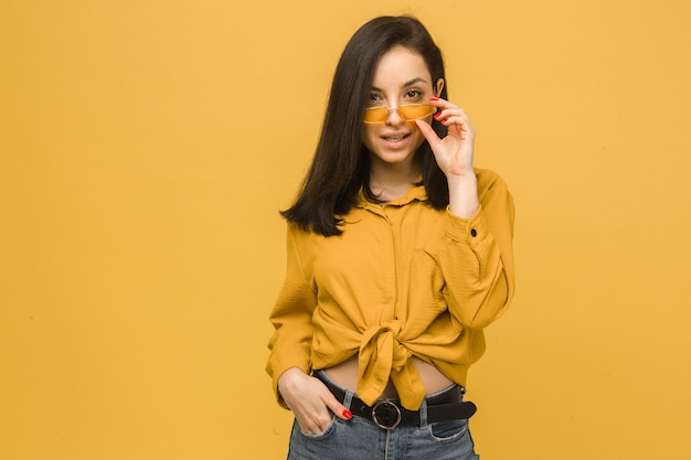 Photo of young female wears sunglasses and looks sexy. Wears yellow shirt, isolated yellow color background.