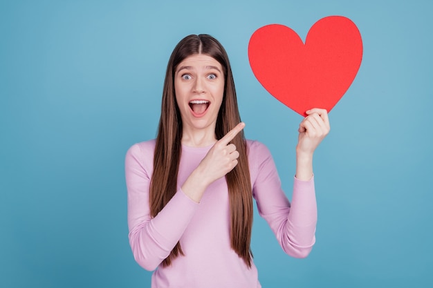 Photo of young girl point finger big paper heart valentine day advert advice isolated over blue color background