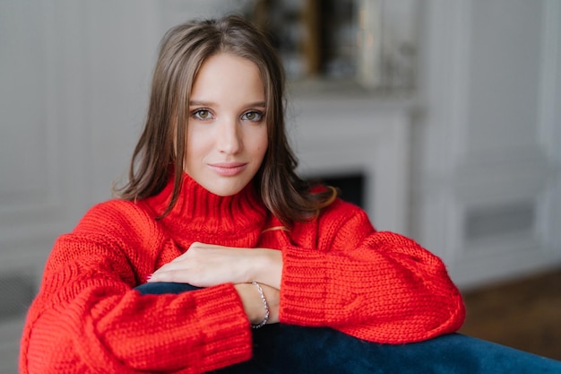 若い格好良いブルネットの女性の写真は、家で休日を楽しんでいますカメラを真剣に見て、ぼやけた背景に対して自宅で暖かいゆるい赤いセーターのポーズを着ています居心地のよさと休息の概念