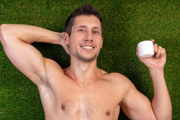 Photo of young handsome naked man laying at the grass with happy smile and enjoying of moisturizing his skin