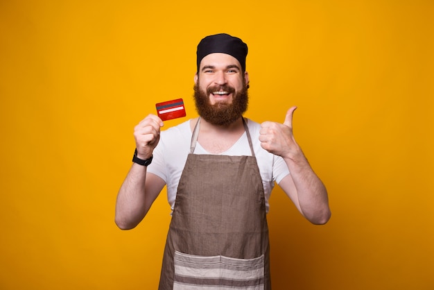 Foto di giovane cuoco unico felice che mostra la carta di credito rossa mentre controllando