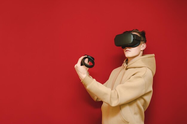 Photo of young man in bright hoodie standing with controllers in hands on red background and playing games on vr helmet looking away vr gaming helmet concept copy space