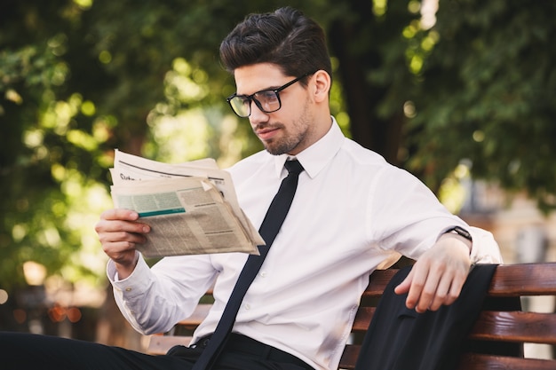 Photo of young man in businesslike suit sitting on bench in green park, and reading newspaper during sunny day