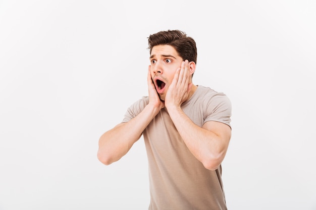 Photo of young man in horror grabbing his face and screaming while looking aside, isolated over white wall