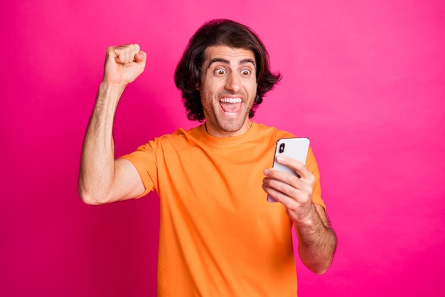 Photo of young man open mouth hold cellphone look screen fist up wear orange t-shirt isolated pink color background