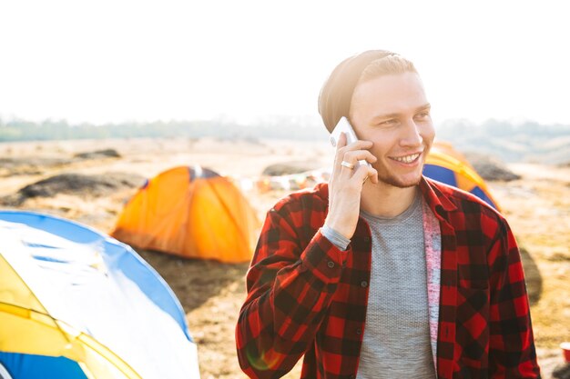 Photo of young man outside in free alternative vacation camping over mountains talking by mobile phone.