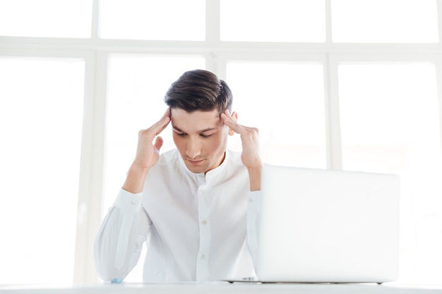 Photo of young man with headache dressed in white shirt using laptop computer. Coworking. Eyes closed.