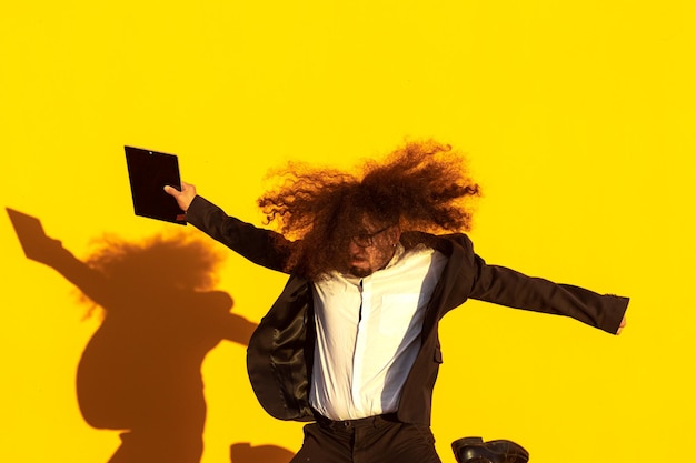 Photo of a young man with long hair jumping with a digital tablet on yellow background