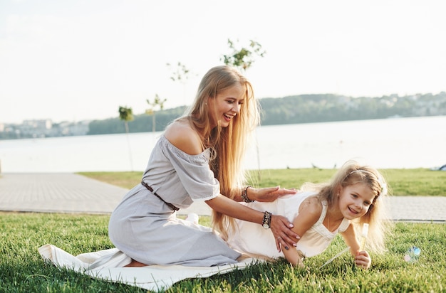 Photo of young mother and her daughter having good time on the green grass with lake at background.