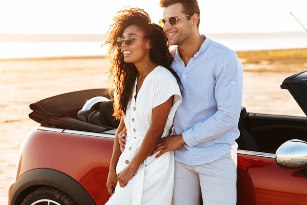 Photo of young multiethnic couple man and woman hugging and smiling together while standing by car outdoors