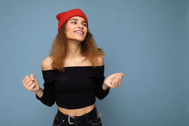 Photo of young positive happy attractive brunette curly woman with sincere emotions wearing black crop top and red hat isolated on blue background with empty space and celebrating victory.