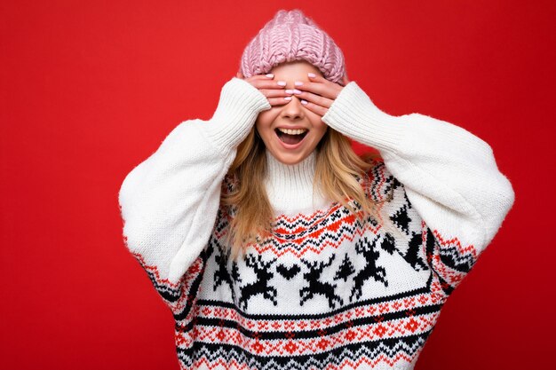 Photo photo of young positive happy beautiful blonde lady with sincere emotions wearing pink knitted hat and winter pullover isolated over red background with empty space and covering eyes with hands.