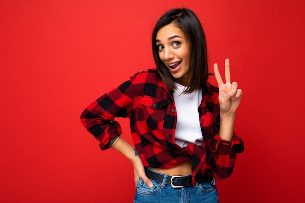 Photo of young positive happy smiling beautiful woman with sincere emotions wearing stylish clothes isolated over background with copy space and showing peace gesture