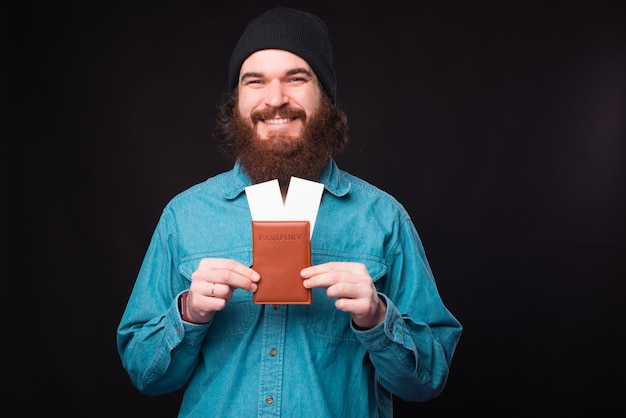 A photo of a young positive man holding a passport and some flying tickets in it near a black wall