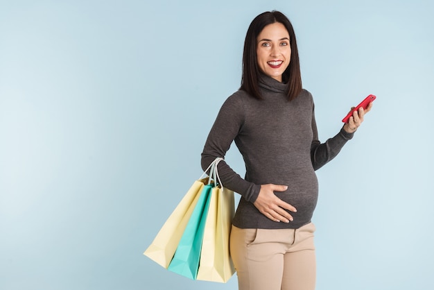 Photo of a young pregnant woman isolated using mobile phone holding shopping bags.