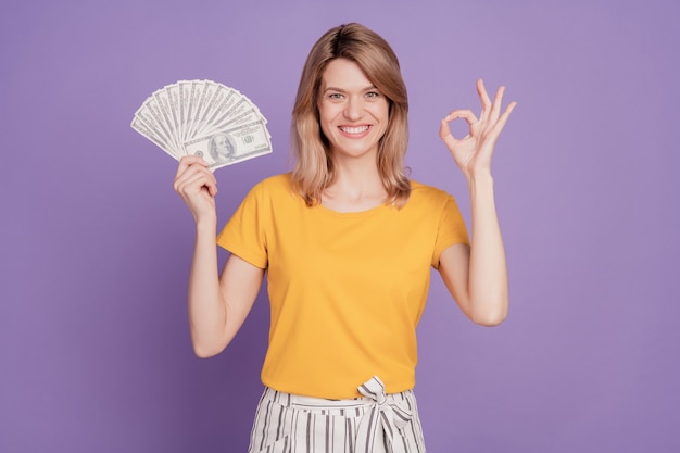 Фотография молодой красивой женщины показывает хорошо, круто, знак доллара, валюты, богатой, изолированной на фиолетовом цветном фоне