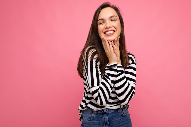 Photo of young smiling positive happy pretty brunette woman with sincere emotions wearing casual striped pullover isolated on pink background with copy space.