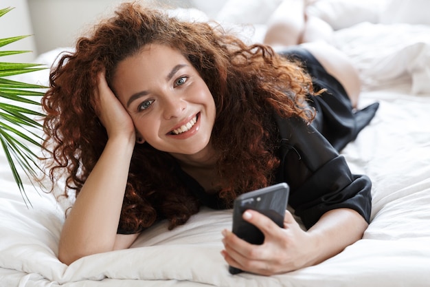 란제리 실크 가운을 입고 웃고 있는 긍정적인 젊은 여성의 사진은 휴대 전화를 사용하여 침대에 있는 홈 호텔에 있습니다.