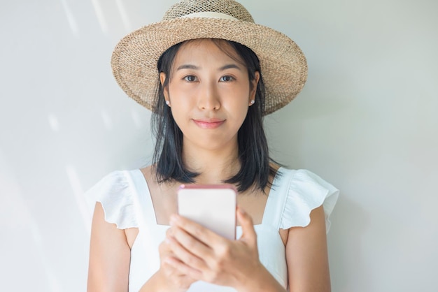 Фотография молодой женщины, счастливой в белом платье и соломенной шляпе, с позитивной улыбкой, использующей смартфон Концепция социальных технологий, путешествия, отдых