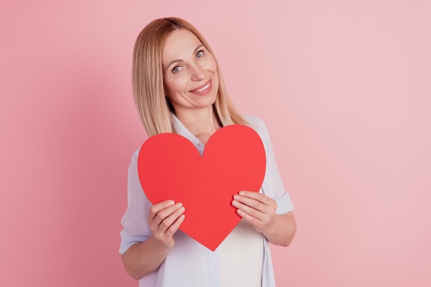 Photo of young woman holding in hand large big heart valentine day card gift isolated over pink background