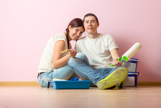 Photo of young woman and man with paint roller sitting on floor