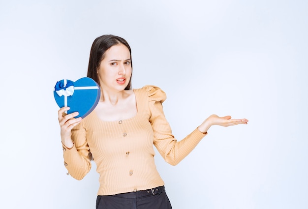 Photo of a young woman model holding a heart shaped gift box against white wall.