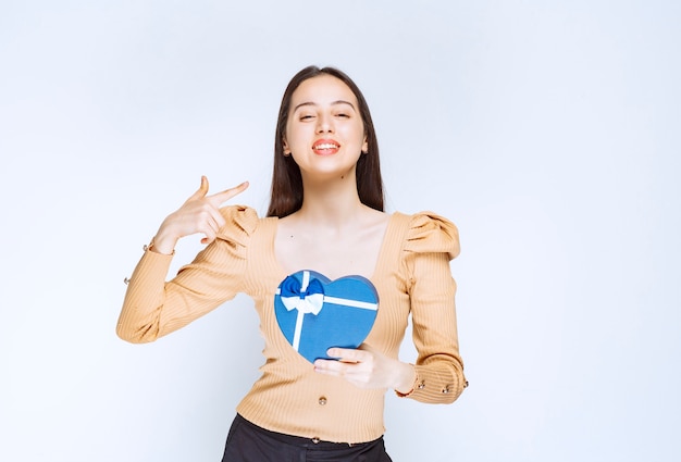 Photo of a young woman model pointing at a heart shaped gift box against white wall.