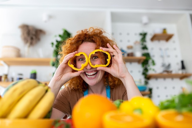 Фотография молодой женщины, улыбающейся и держащей круги перца на глазах во время приготовления салата со свежими овощами в кухонном интерьере дома