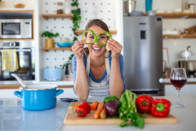 Photo of young woman smiling with green pepper slices while cooking salad with fresh vegetables in kitchen interior at home