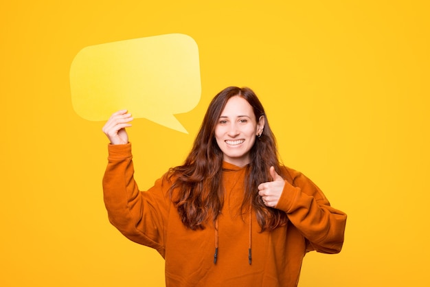 Photo of a young woman that is holding a speech bubble showing a thumb up is smiling