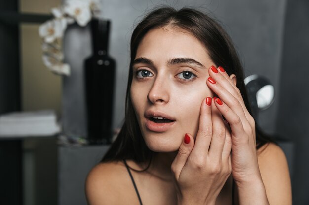 Photo of young woman with long dark hair and clean skin looking\
ahead and touching her face