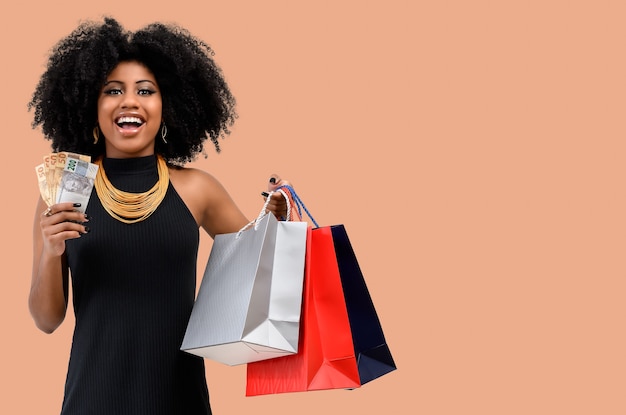 Фотография улыбающейся молодой афро-женщины, держащей сумку с покупками и бразильских денежных купюр